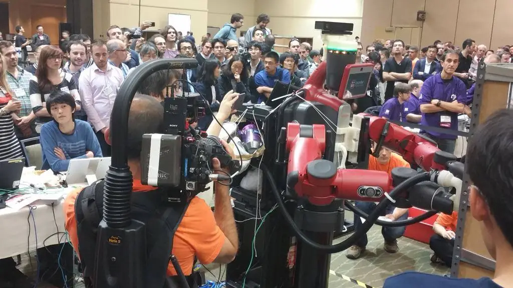 A crowd watching a red two-armed robot pick items at the Amazon Picking Challenge.